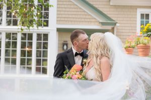 Bride and groom share a romantic kiss outdoors, bride holding colorful bouquet with veil flowing, elegant venue backdrop.