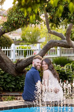 Couple smiling by a fountain under a tree in a garden with white fence At Prescott Park Portsmouth New Hampshire, engagement photo session.