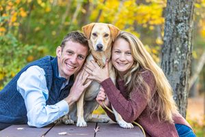 Smiling couple posing with their yellow Labrador dog during fall engagement photos. Ellacoya State Park Gilford NH