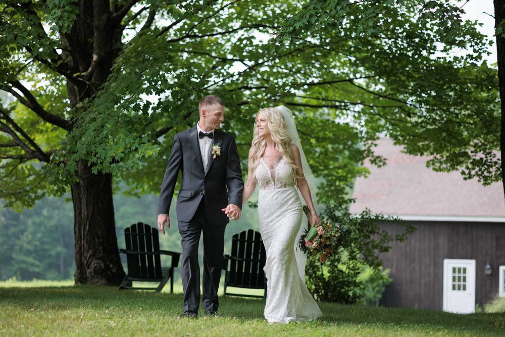 Bride and groom walk hand-in-hand beneath large shade tree on lush green lawn at outdoor wedding.