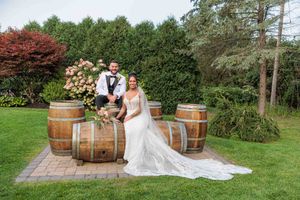 Bride and groom posing together among rustic wooden wine barrels in a lush garden setting on their wedding day.