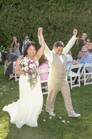 Newlywed couple celebrating with raised hands after outdoor wedding ceremony on green lawn with guests applauding.