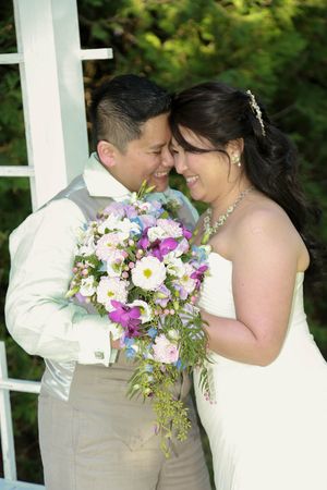 A smiling bride and groom in wedding attire touch foreheads tenderly while holding a colorful pink and purple floral bouquet.