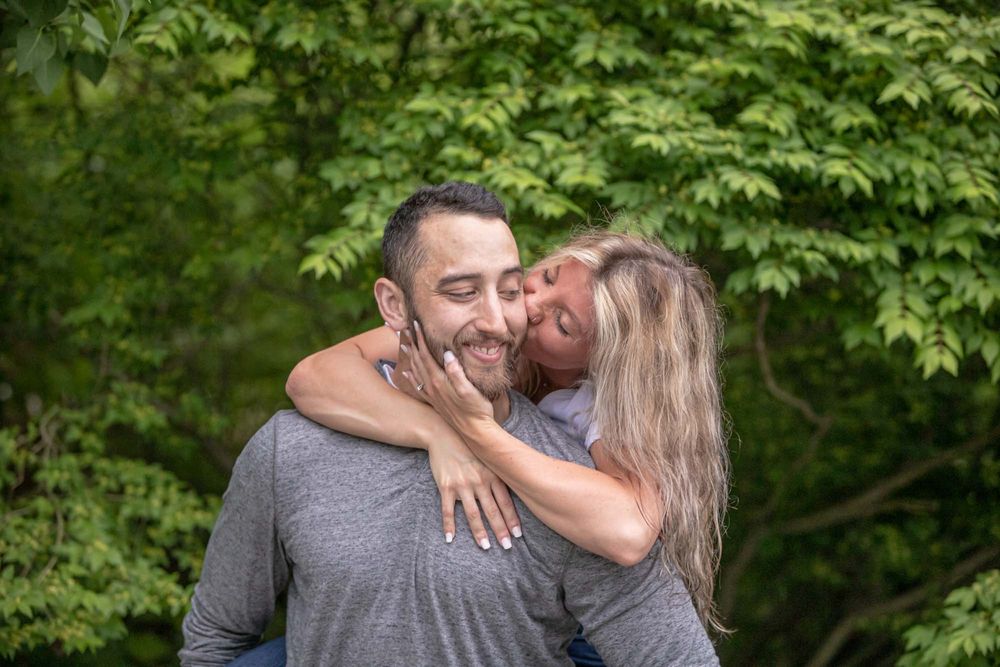 A woman kisses a smiling man on the cheek outdoors, both wearing gray shirts with lush green trees behind them.