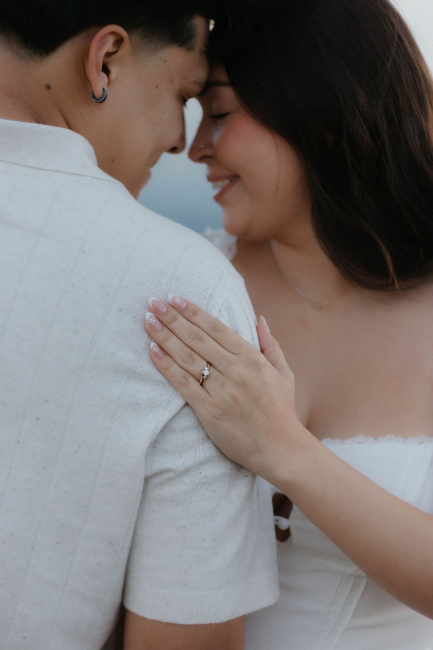 A romantic engagement photoshoot showing a couple embracing while wearing white, with focus on the ring and tender moments.