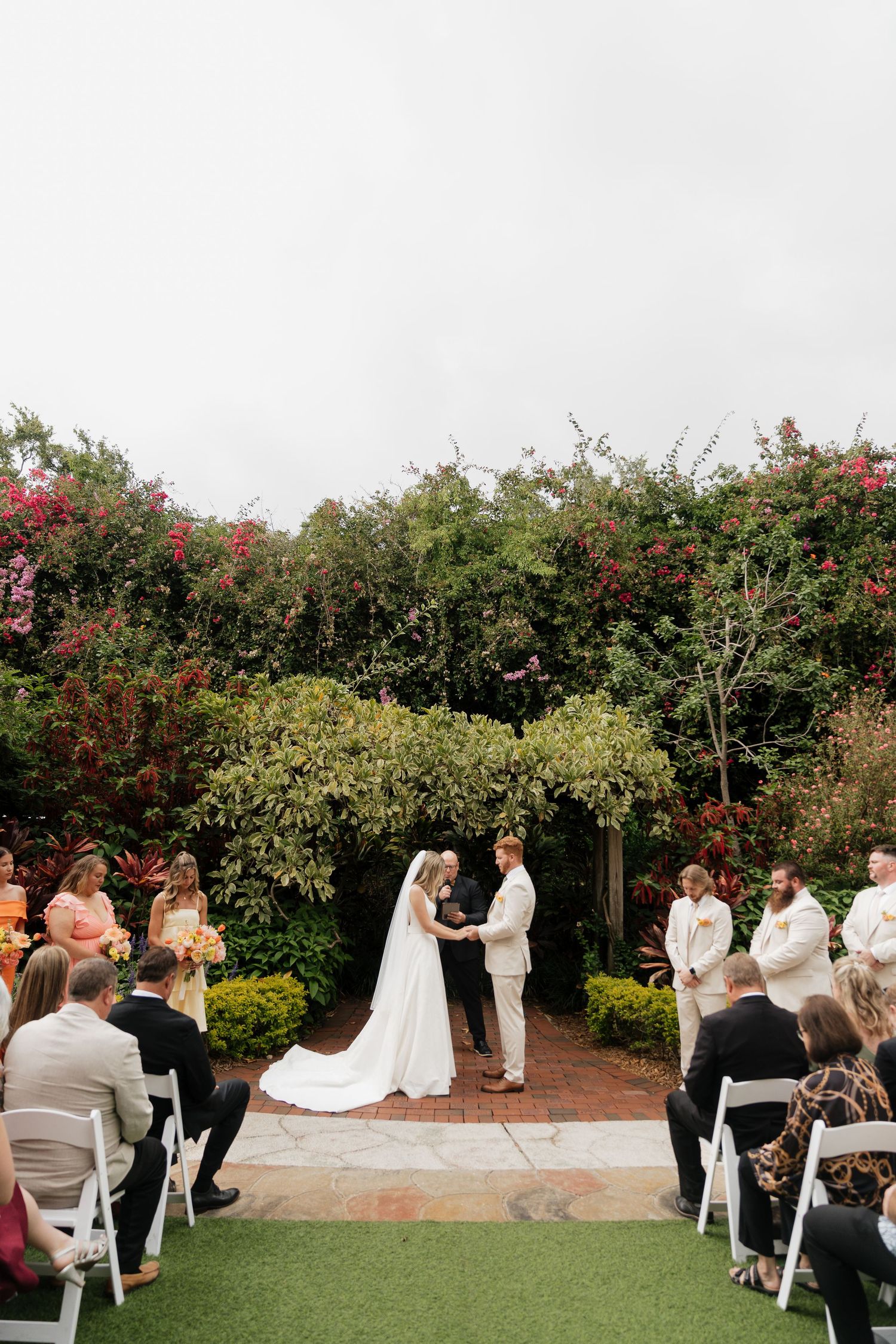 Wedding ceremony guests seated in white chairs on a garden lawn with red flowering trees in the background.