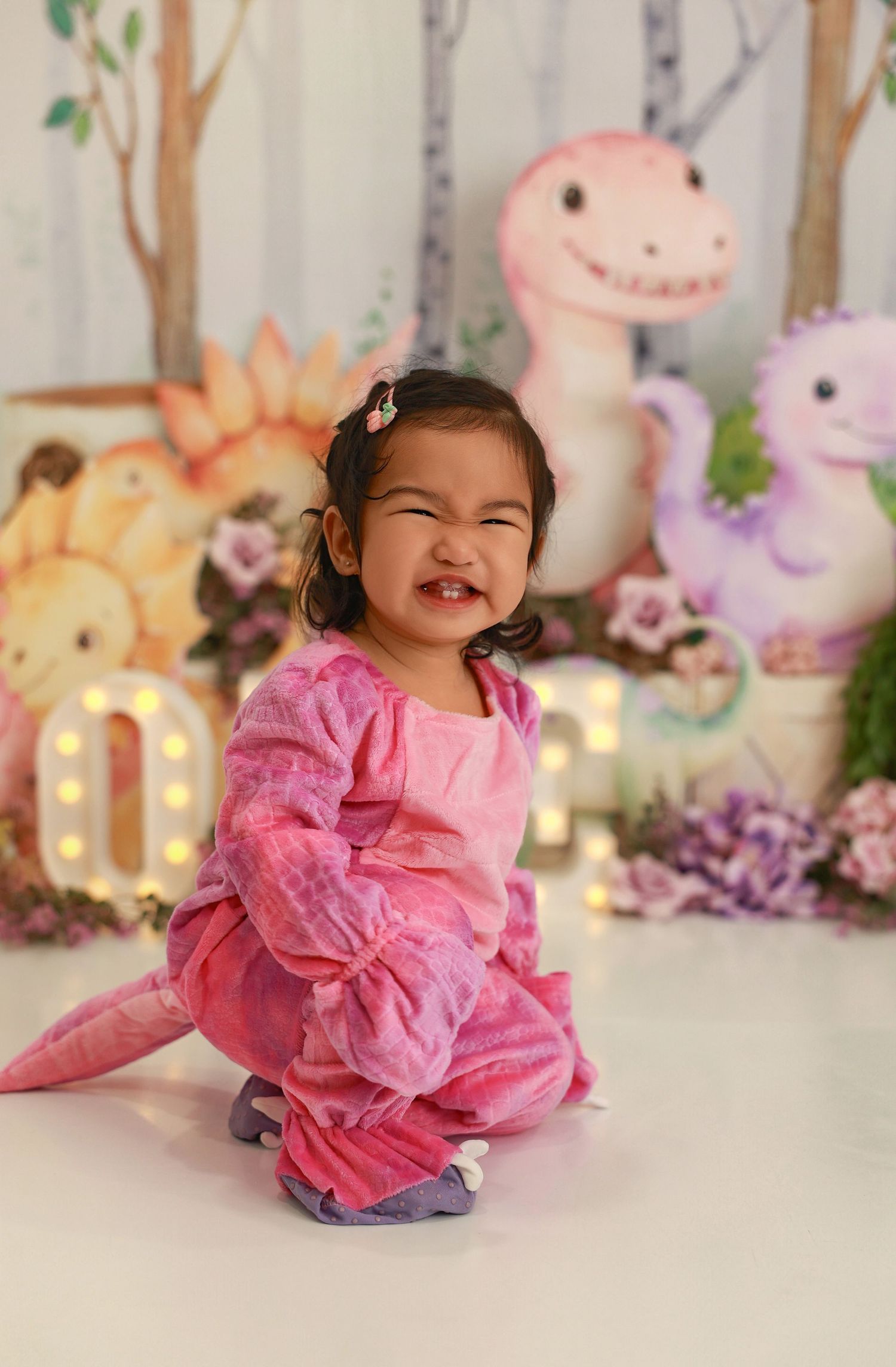 A child in a pink ruffled dress plays and poses on the floor with plush toys and decorative lights in the background.