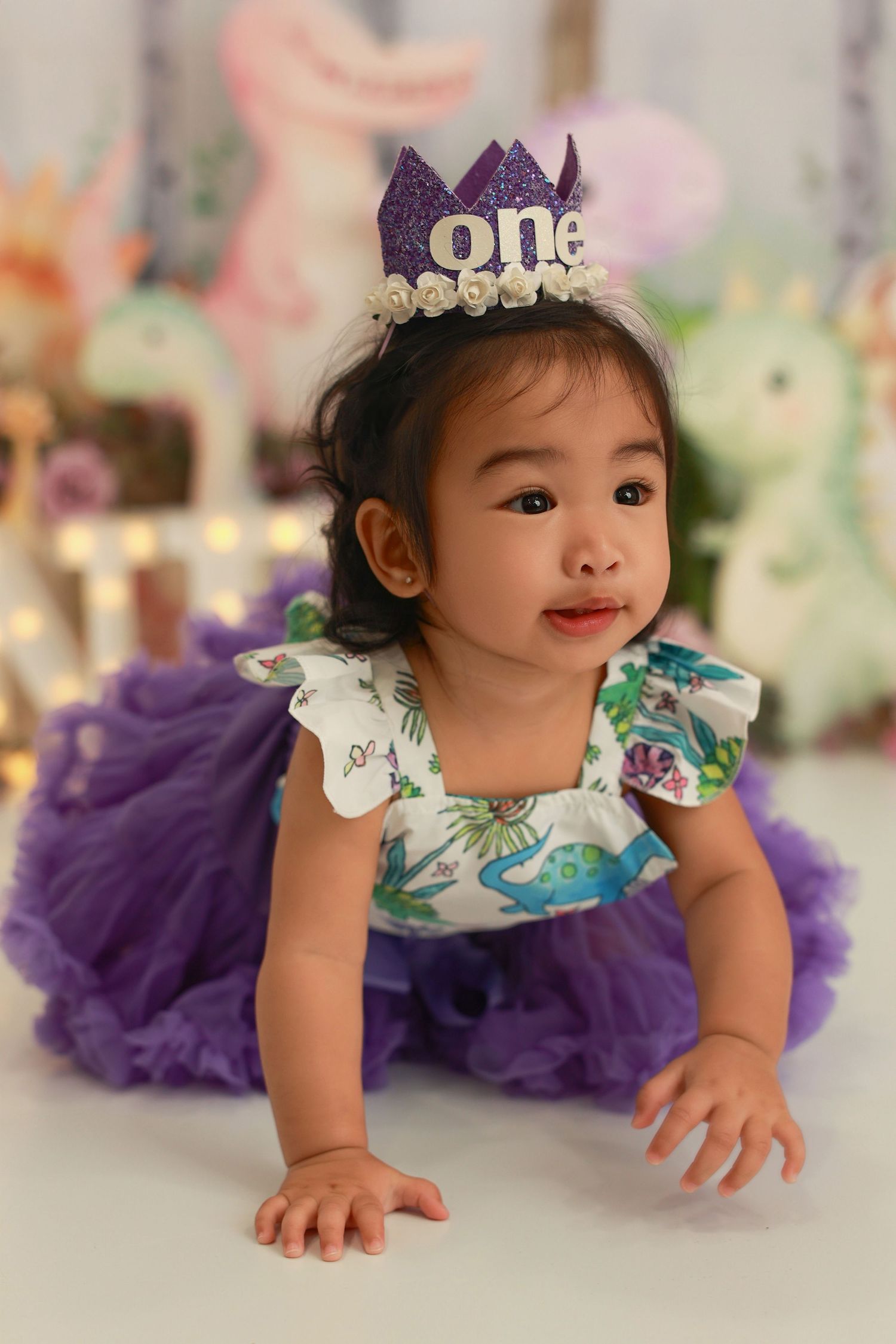 A baby in a floral dress with purple tulle skirt and tiara crawls and poses during a photo session.