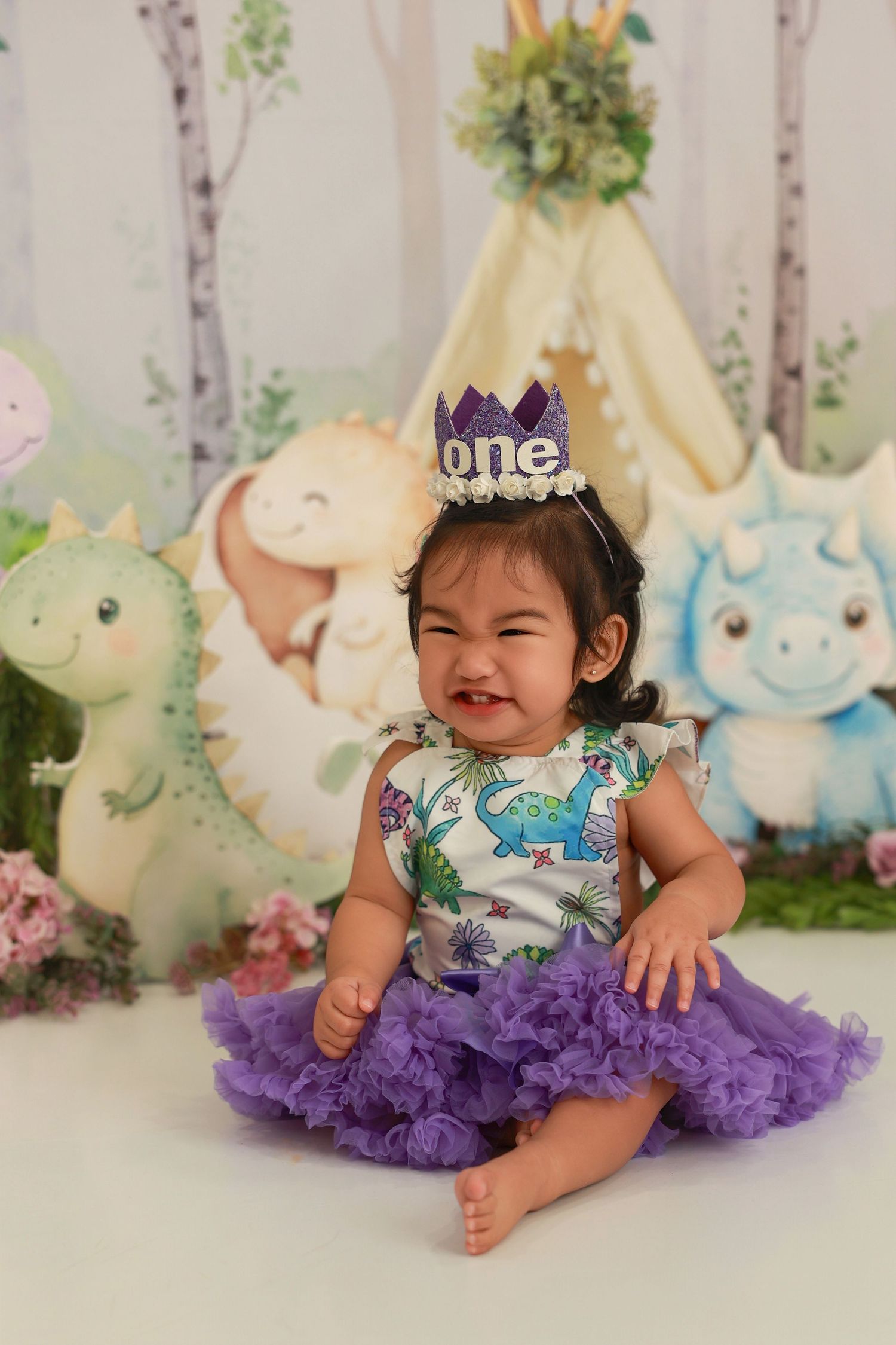 A purple tutu dress and birthday crown create a festive first birthday photo setup with soft plush toys in the background.