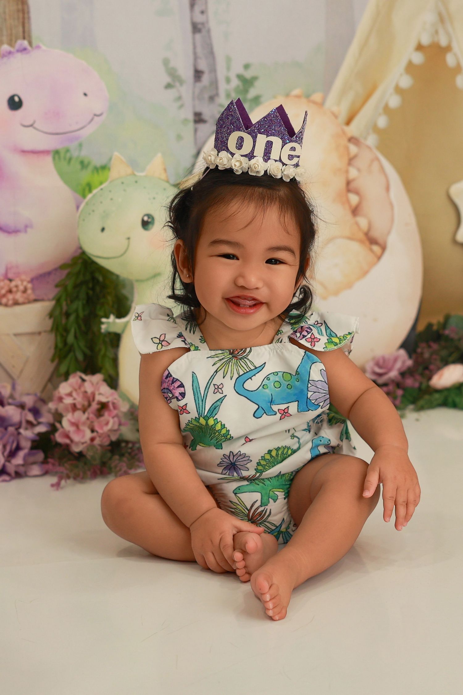 A baby wearing a dinosaur print romper and 'one' crown poses in front of plush dinosaurs and floral decorations.