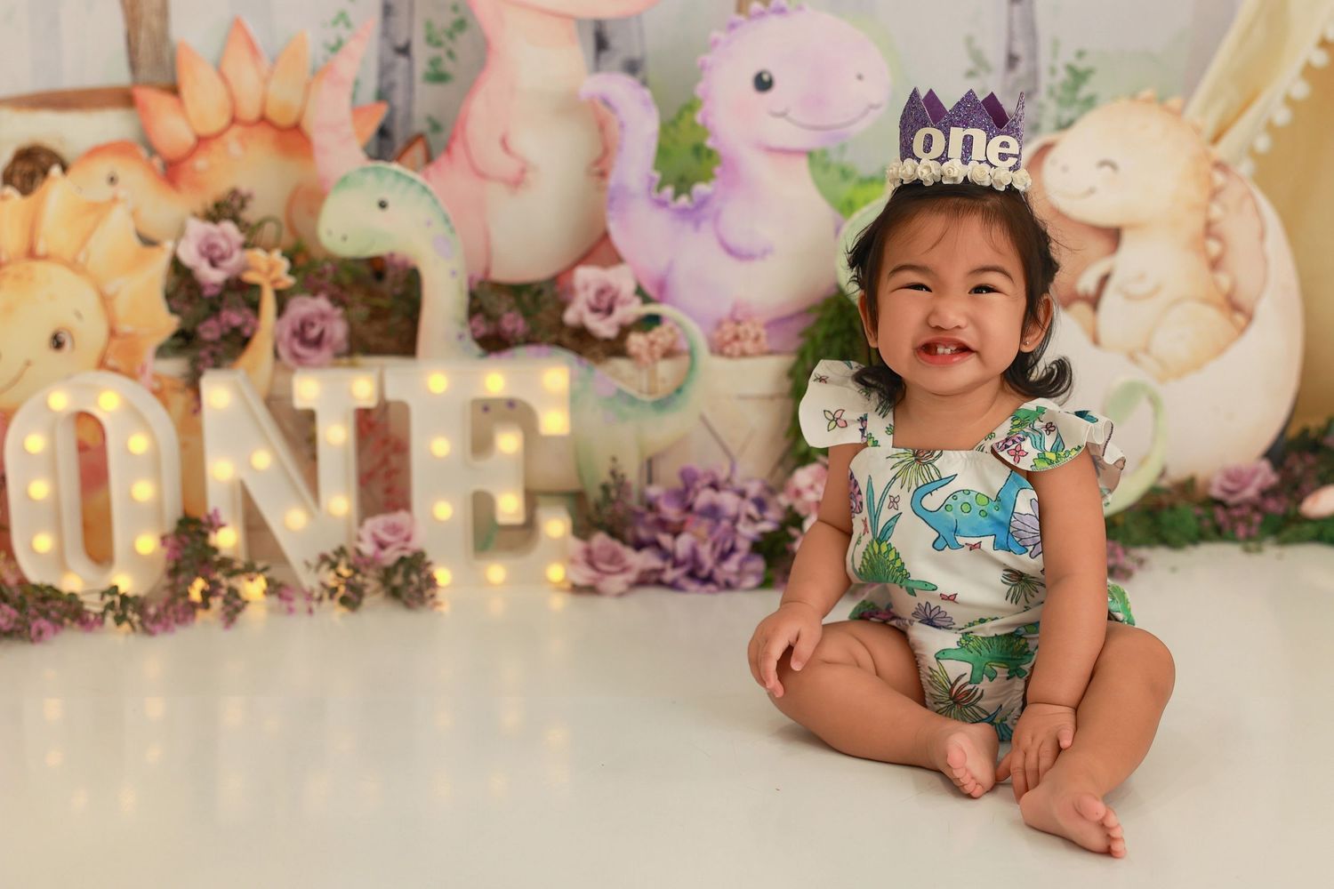 A baby in a floral dress sits in a dreamy garden-themed photo studio setting with pink and cream decorations.