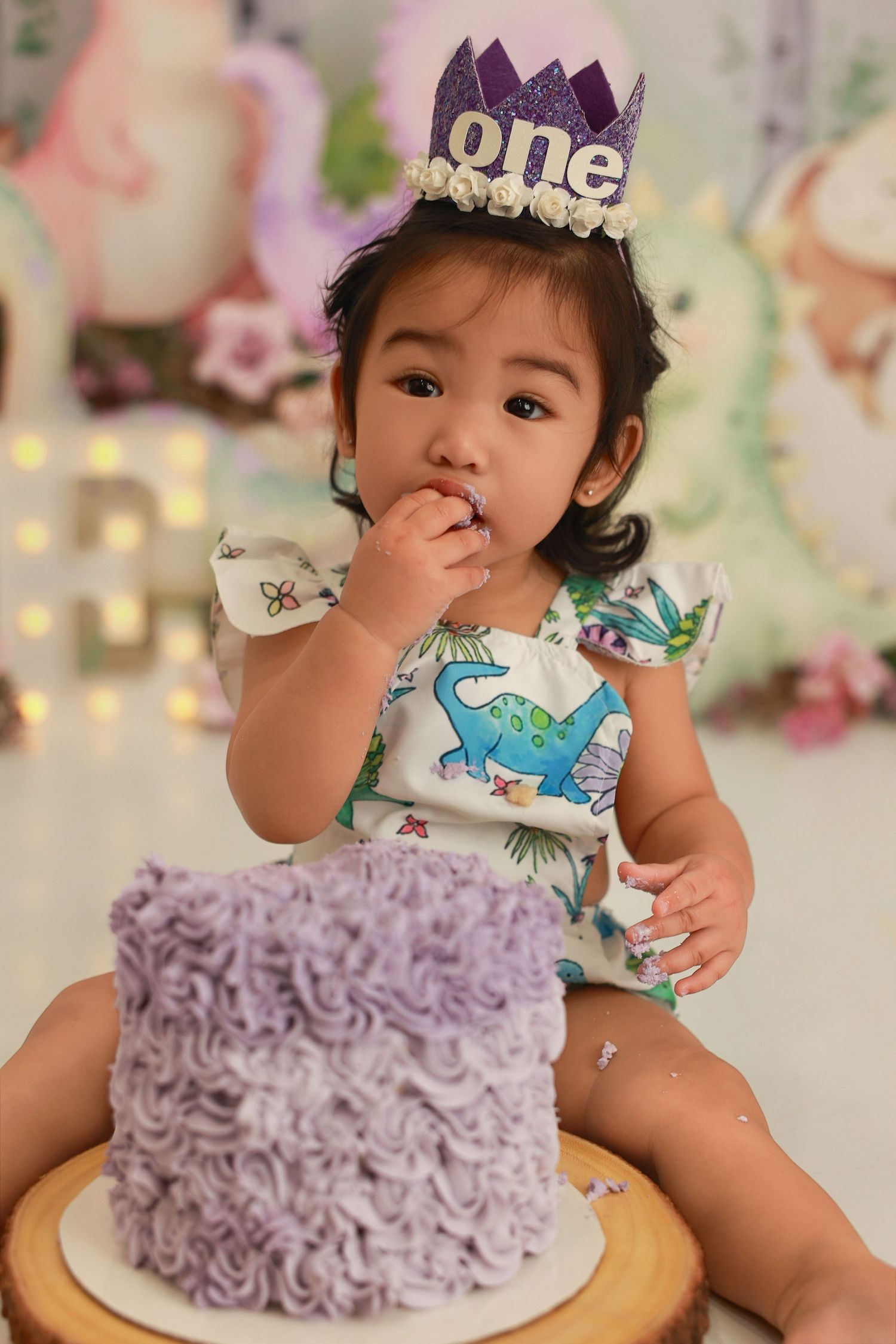 A first birthday celebration scene with a purple frosted cake and decorative party setup against a floral backdrop.