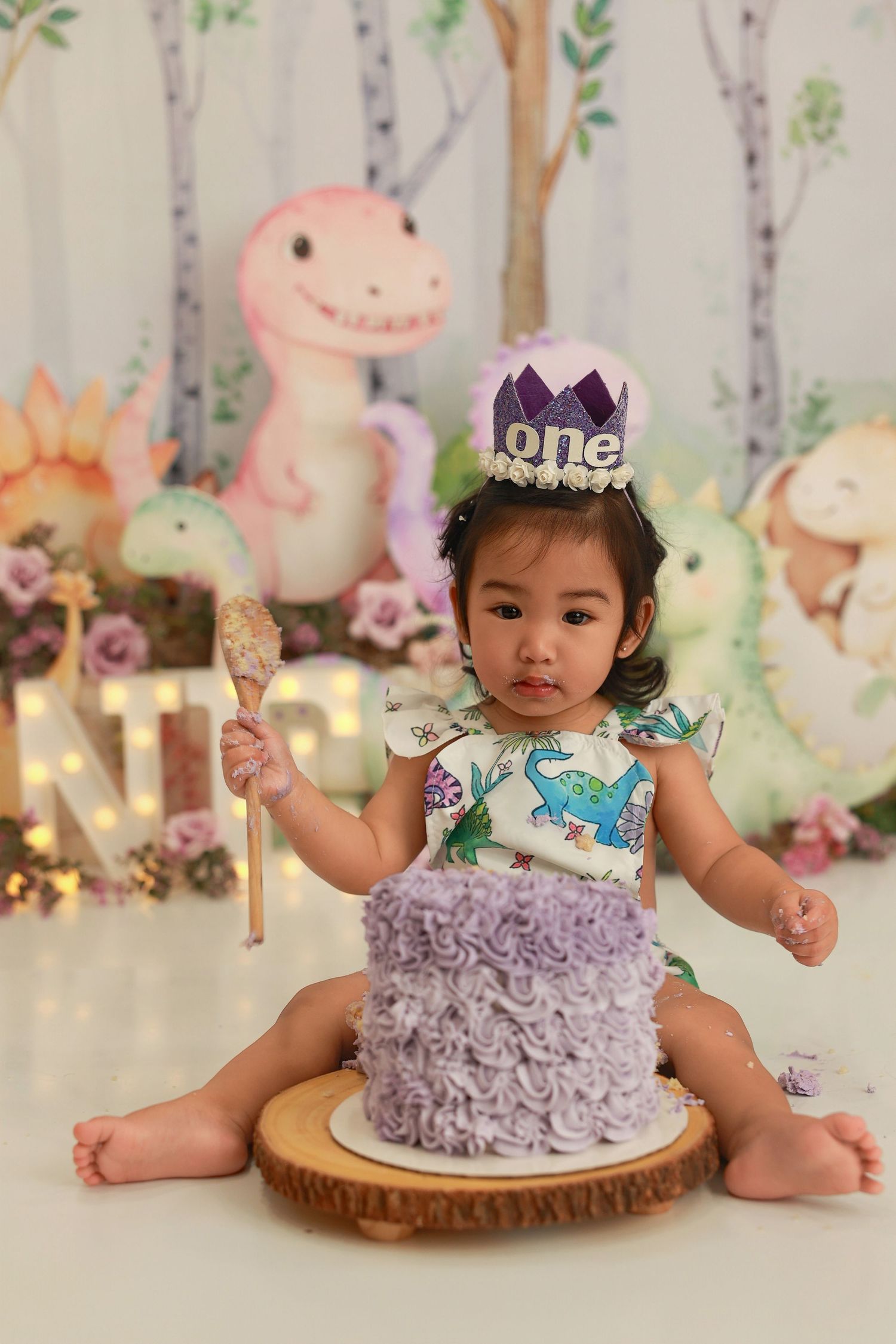 A festive first birthday cake smash setup with a purple ruffled cake and woodland animal decorations in the background.