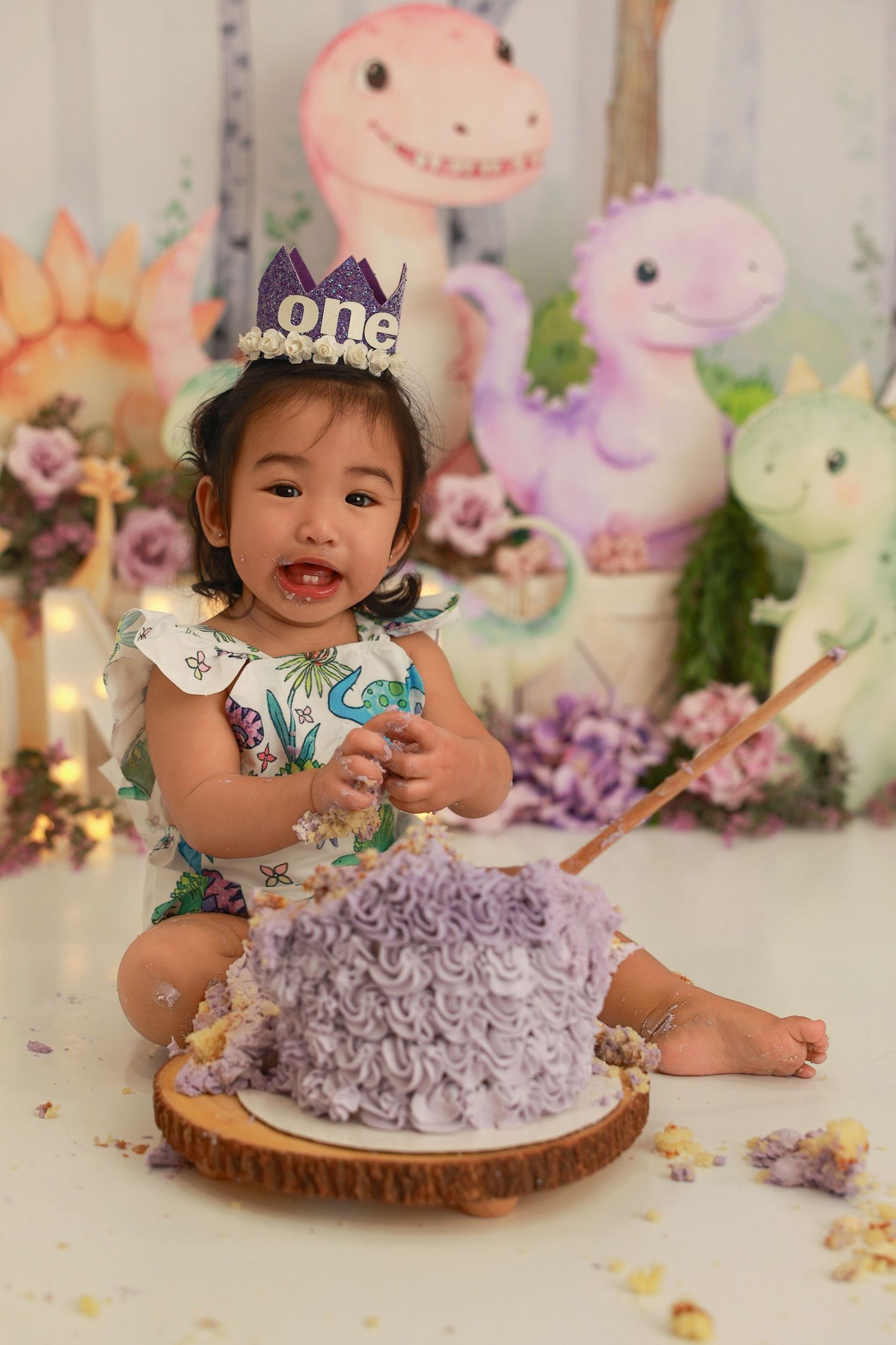 A cake smash photo session featuring a purple frosted birthday cake on a wooden slice with plush animal decor.