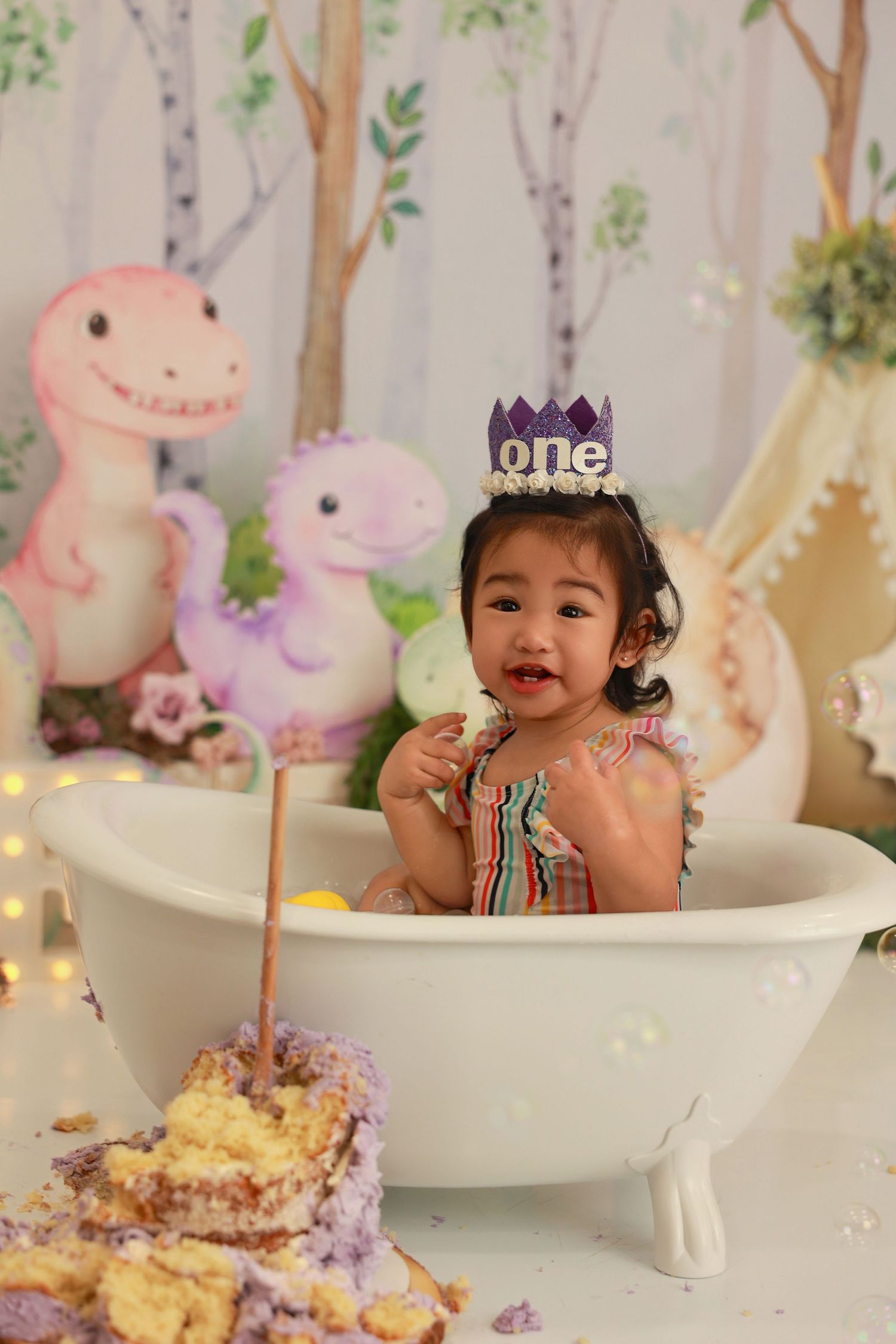 A baby's first birthday cake smash photo session with pink dinosaur decorations and white bathtub in a woodland themed setting.