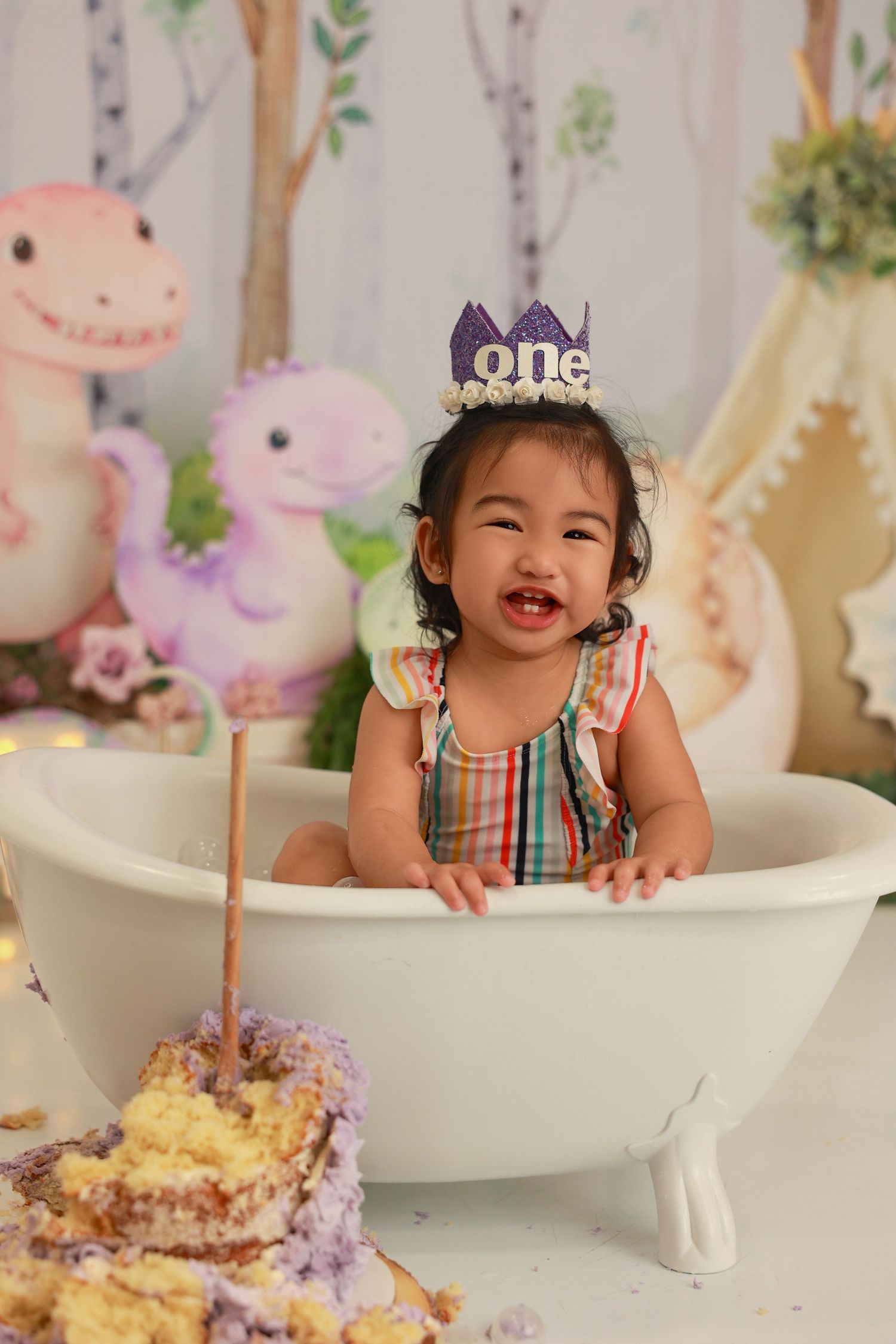 First birthday celebration setup with white bathtub prop, floral decorations, and colorful cake against pastel backdrop.