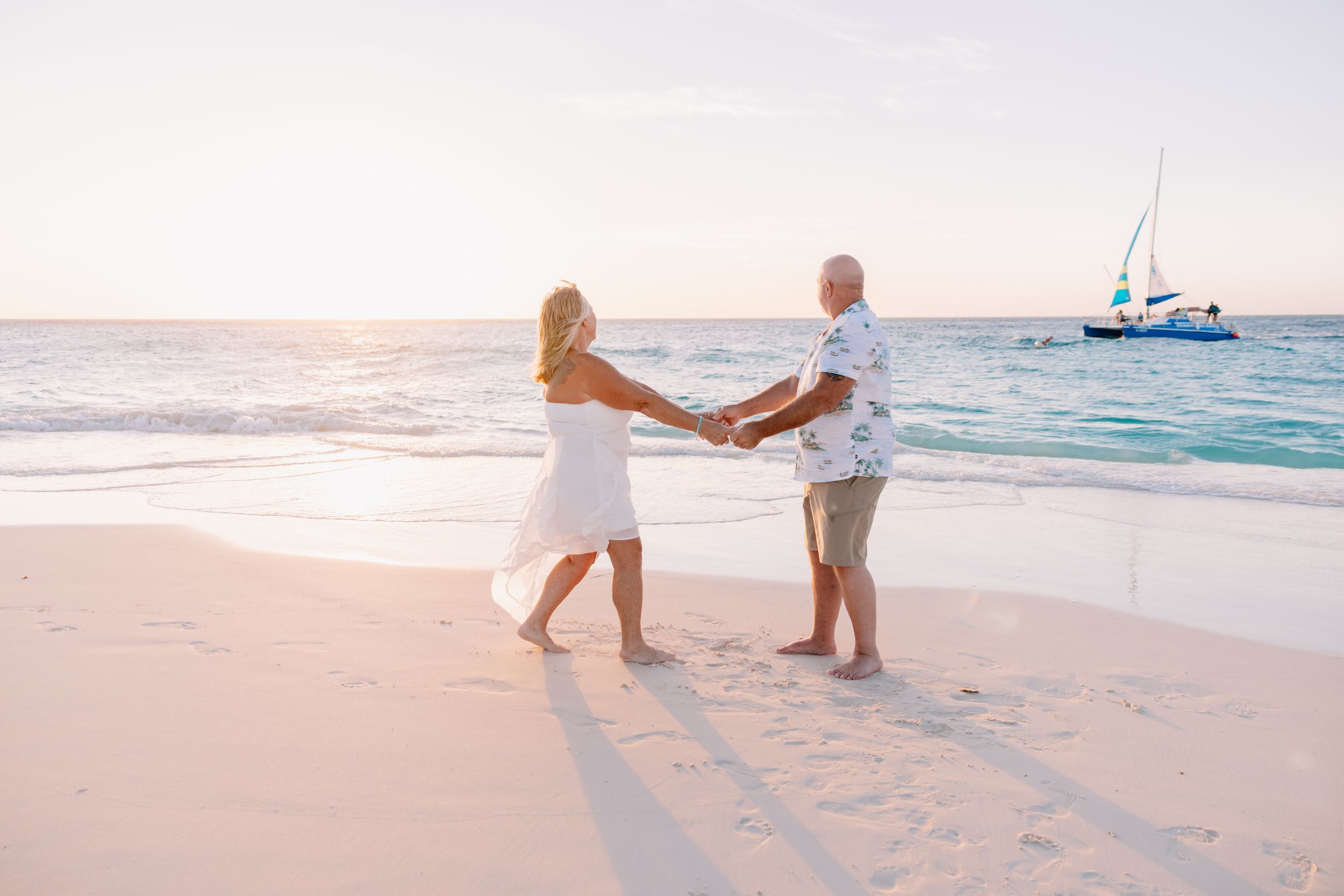 Two people walking hand in hand along a sandy beach with a boat visible in the distance.