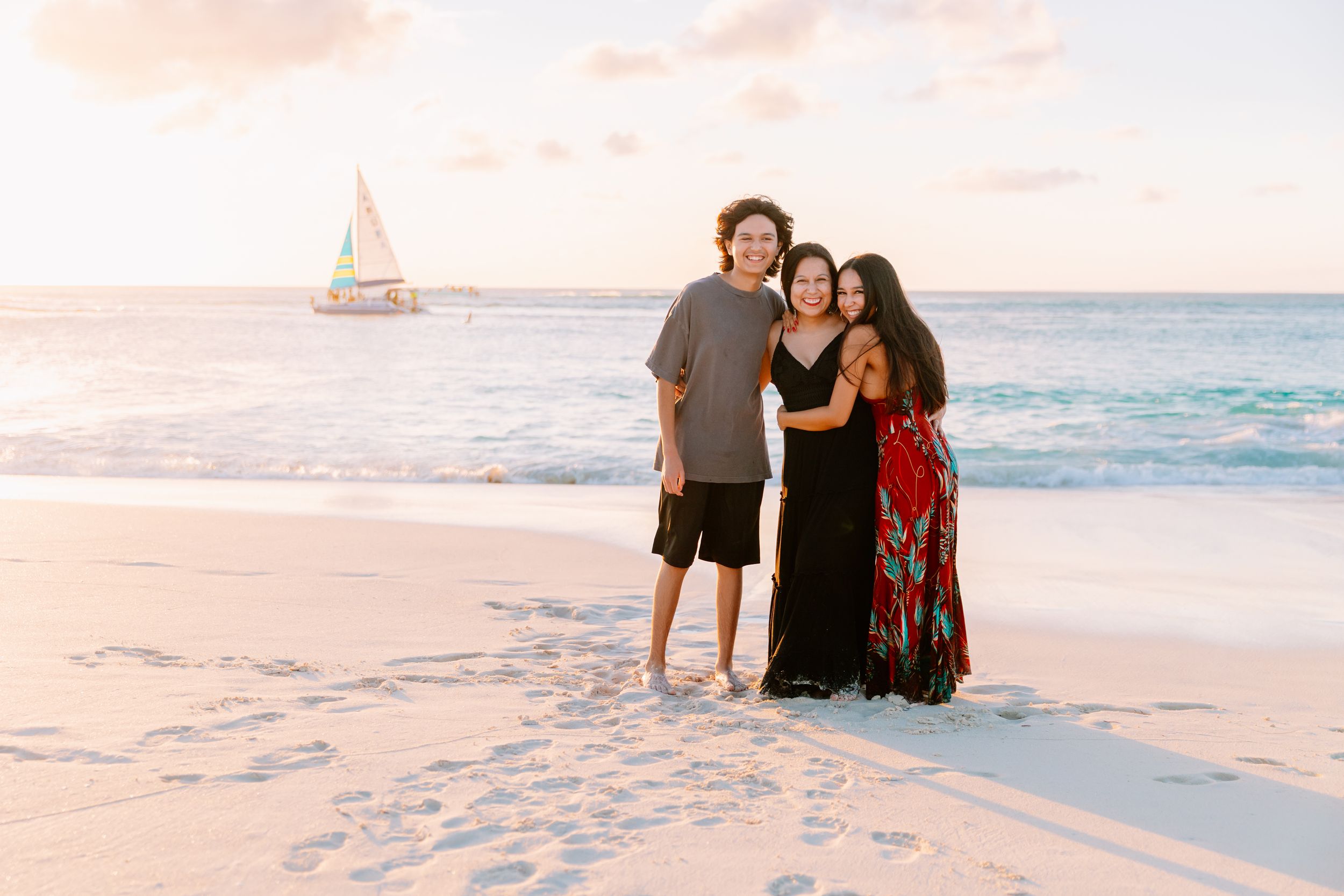 Sunset family photoshoot on pristine beach with sailboat visible in background.