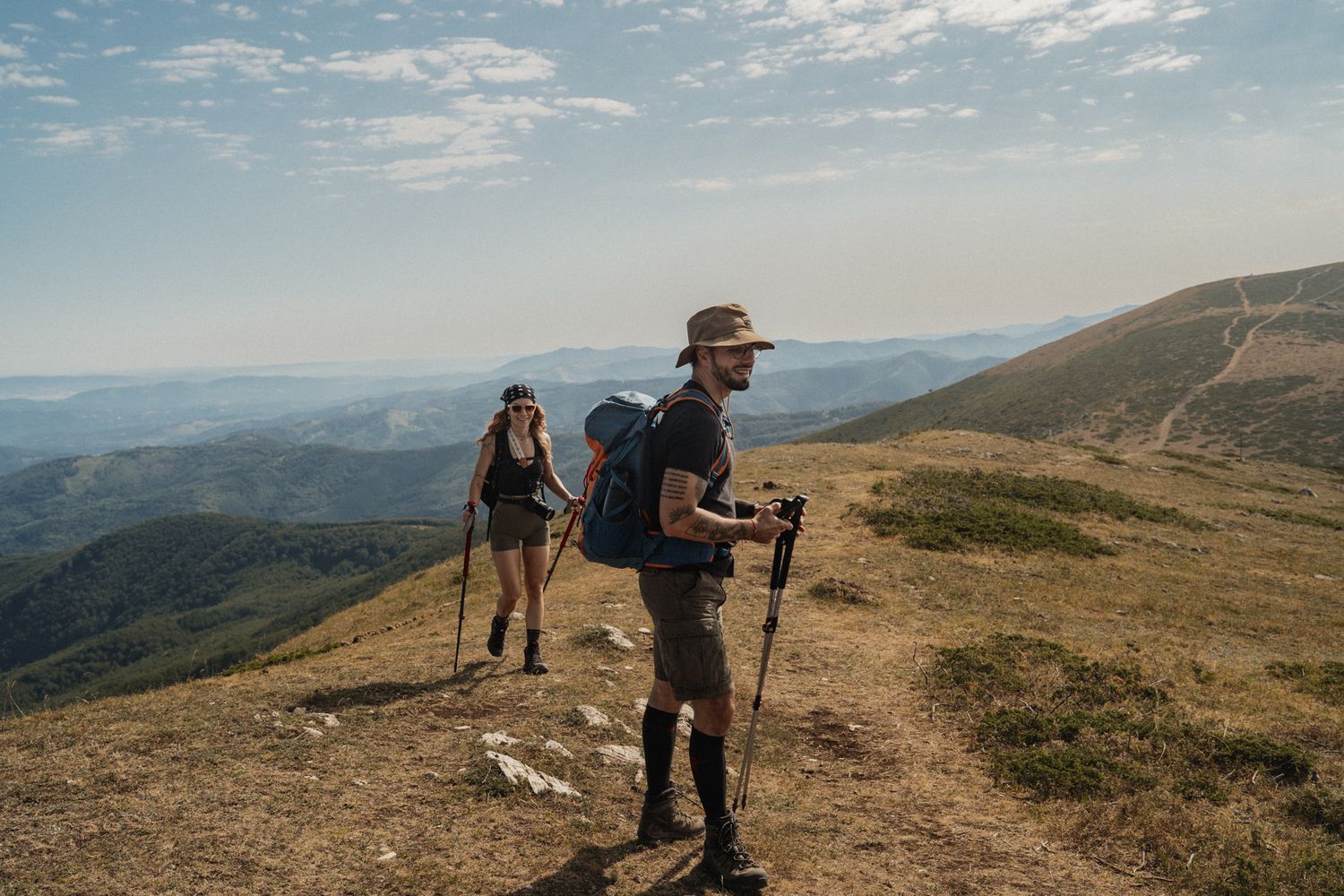 Two hikers traverse a scenic mountain trail with hiking poles and backpacks against a backdrop of rolling hills.