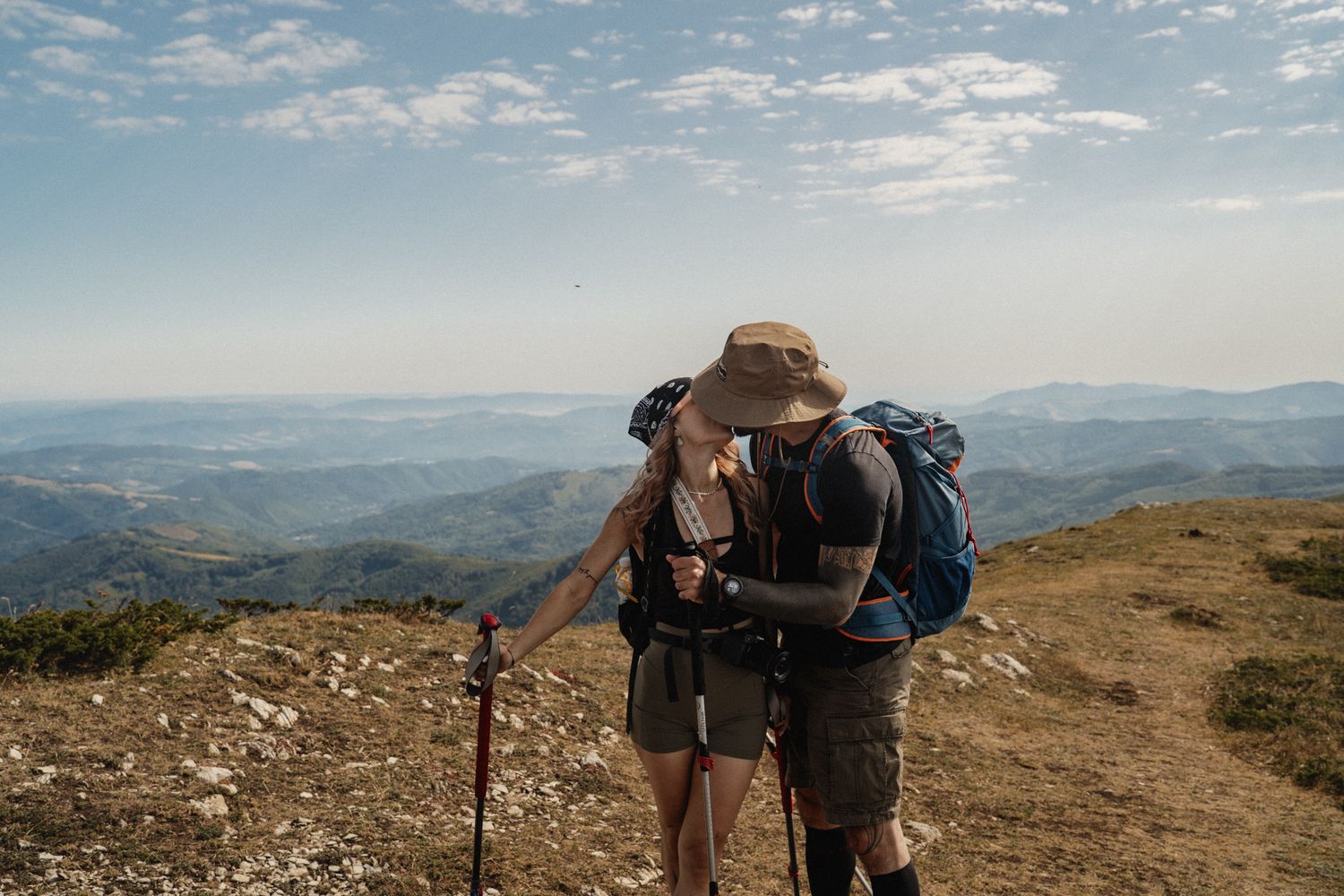 Panoramic view of hikers with backpacks traversing a scenic mountain ridge trail under blue skies and clouds.
