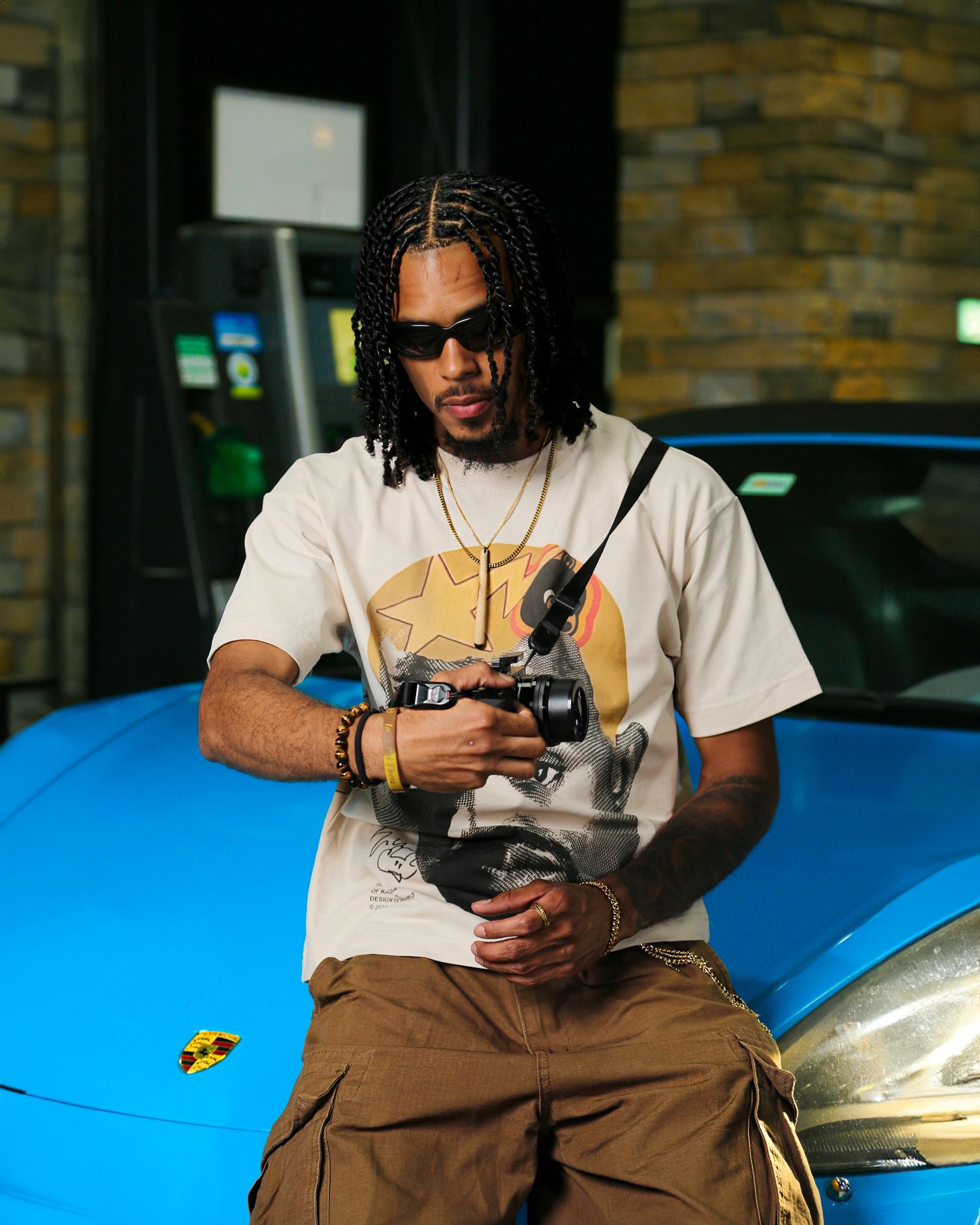 Person in sunglasses and white t-shirt sitting on blue car hood while holding camera equipment.