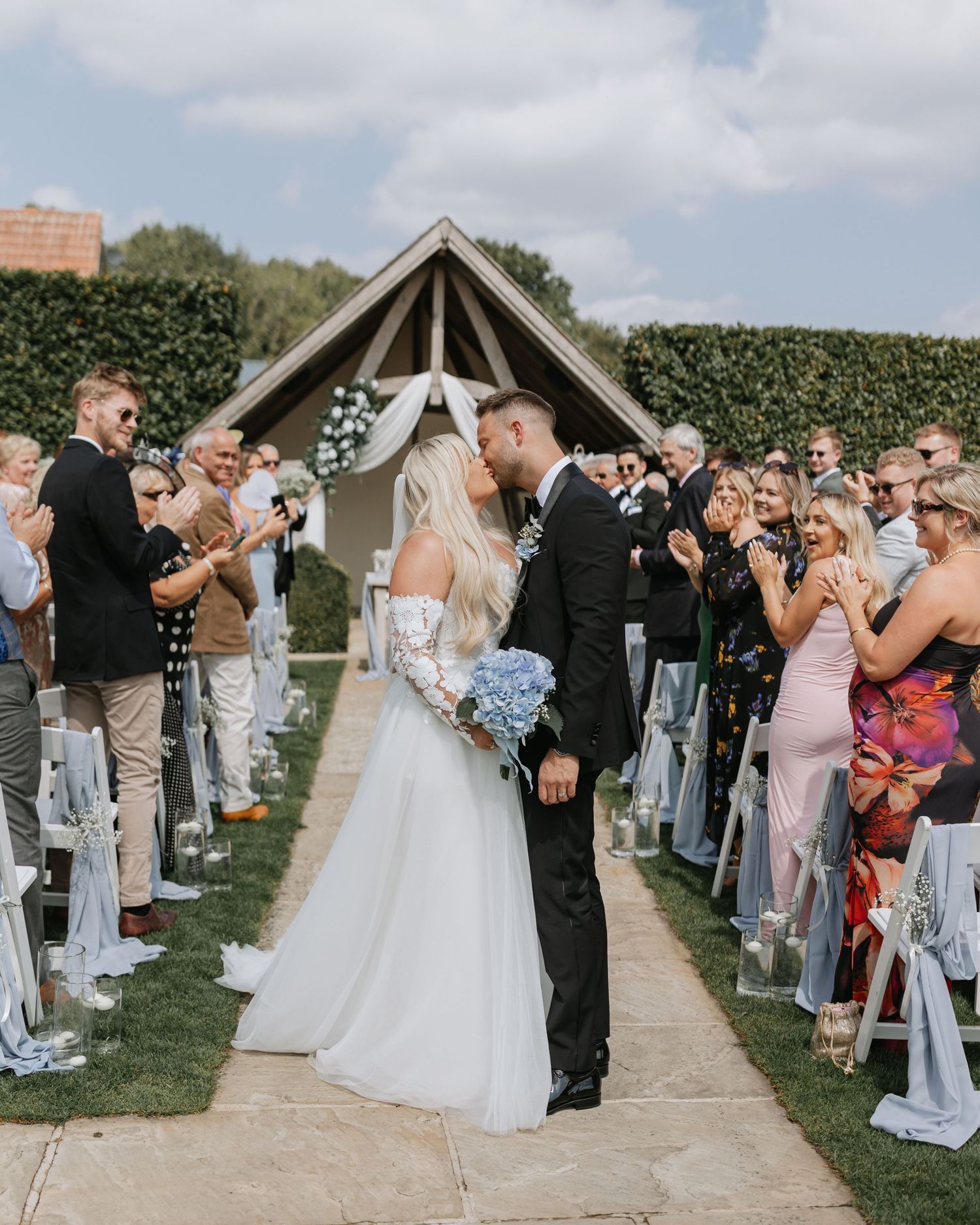 Wedding guests line up outdoors to celebrate as newlyweds share their first kiss during a summer ceremony.