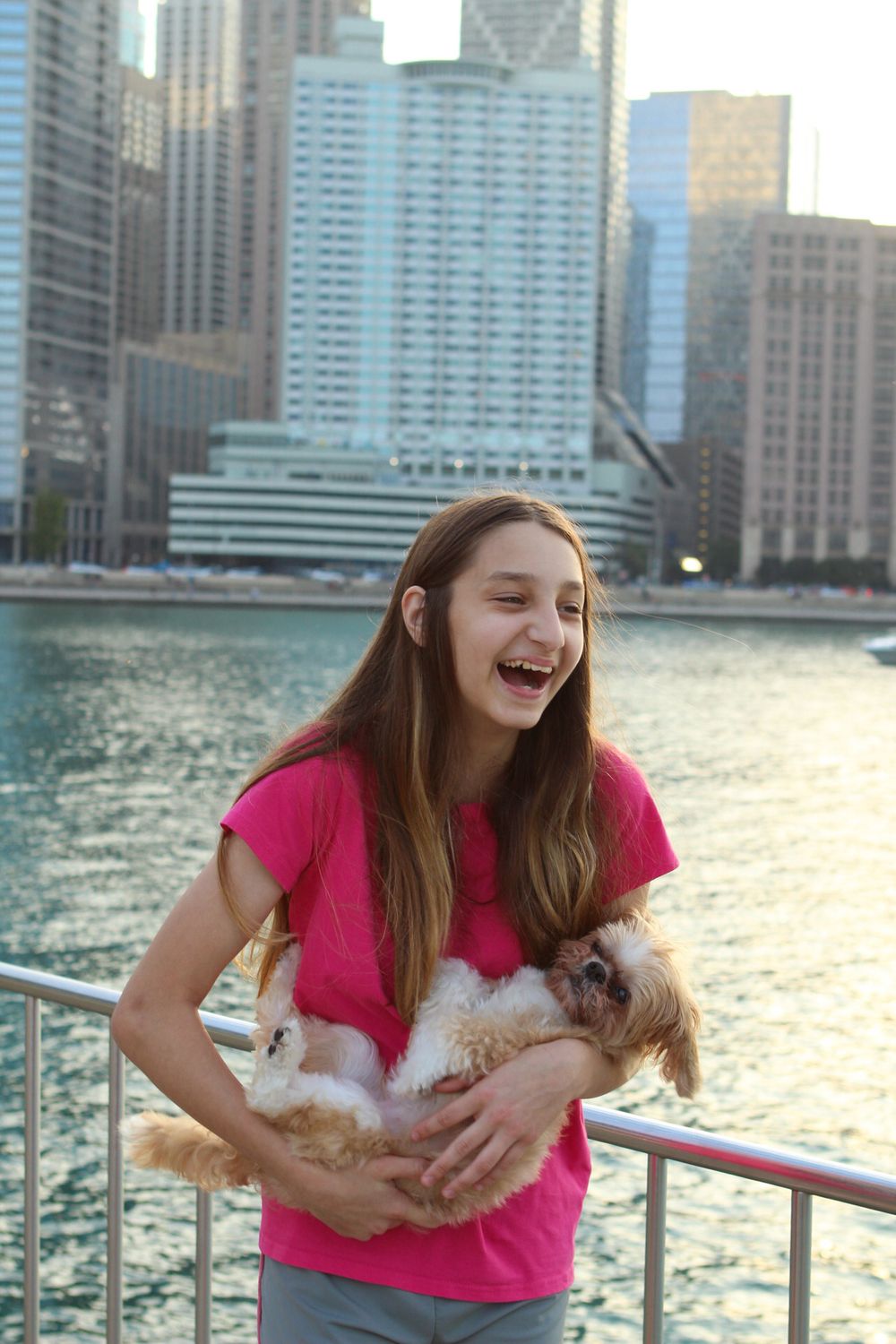 Young person in pink shirt laughing while holding a cat by the waterfront.