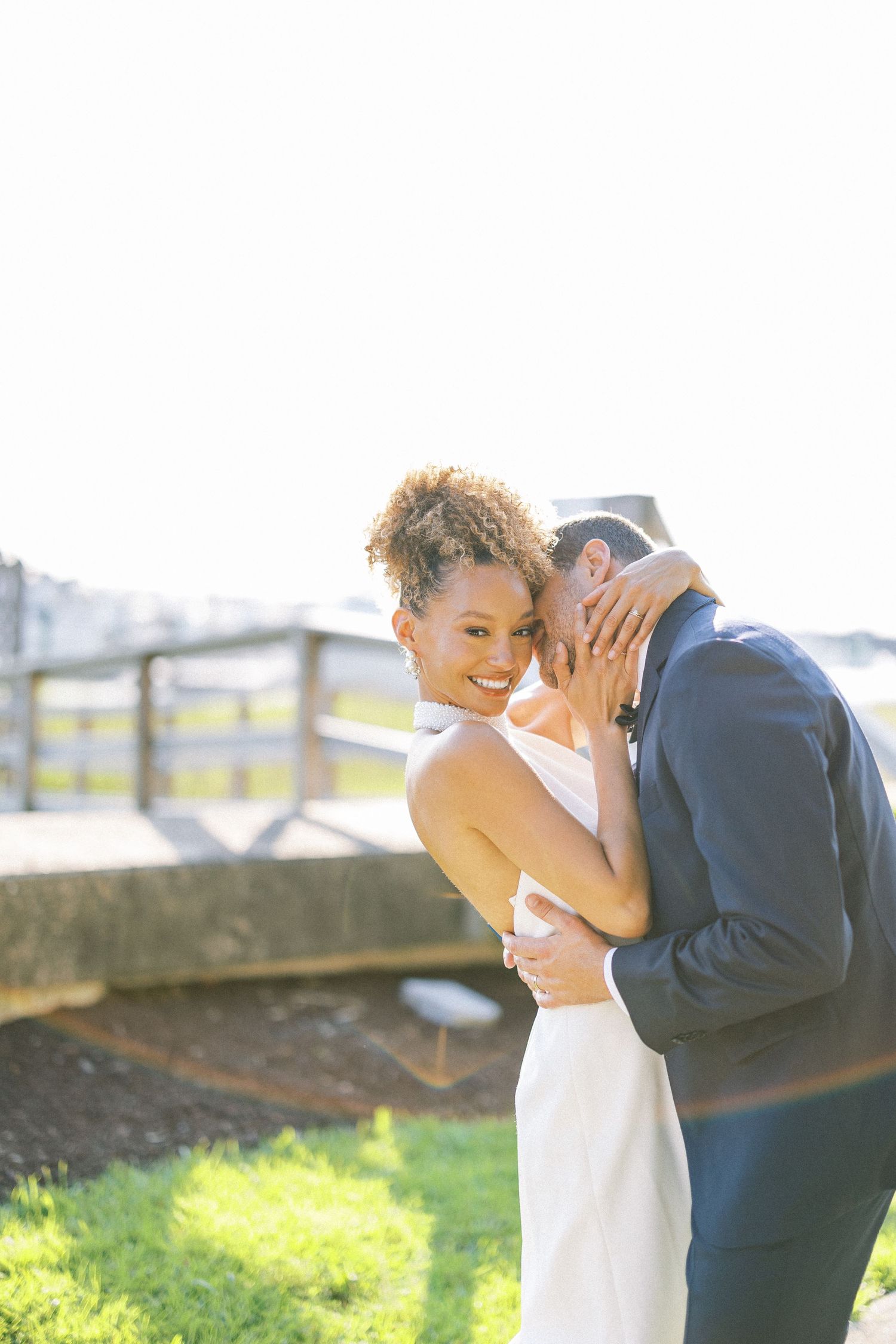 A couple in formal attire embraces tenderly during an outdoor photo session in a sunlit field.