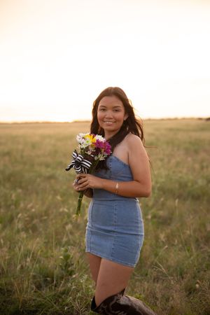 A person in a denim dress holds wildflowers while standing in a sunlit field at golden hour.