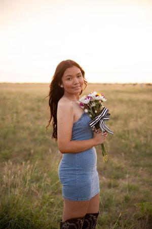 A person in a denim top holds a bouquet of flowers while standing in a sunlit field at golden hour.