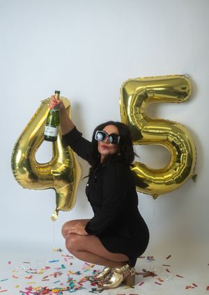 Celebratory scene with large gold foil number balloons 45, champagne bottle, and confetti against white wall.