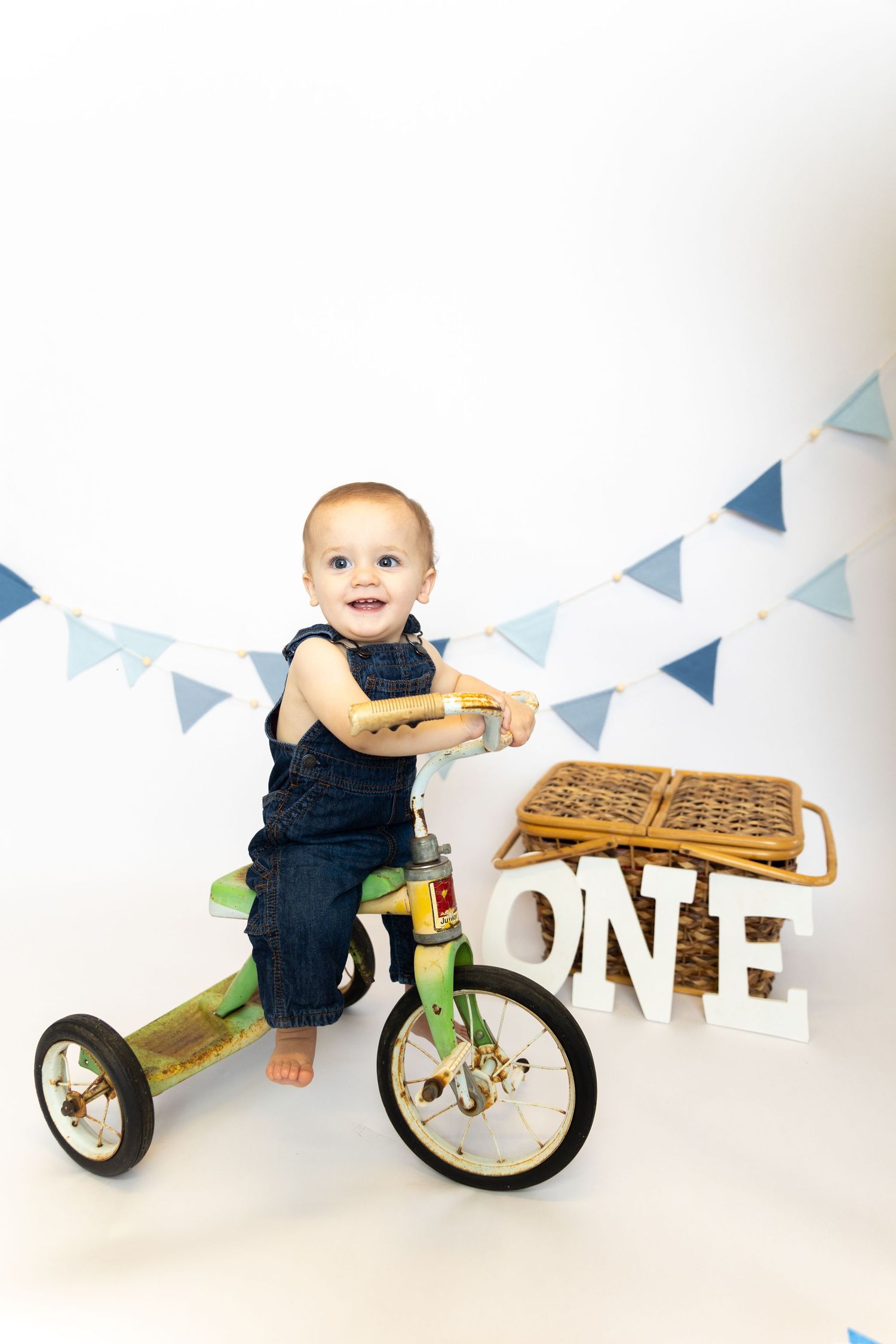 A baby in navy blue overalls plays with a wooden balance bike against a white background with blue bunting.