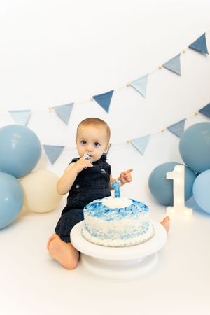 Baby in navy romper explores first birthday cake during cake smash photo session with blue and white party decorations.