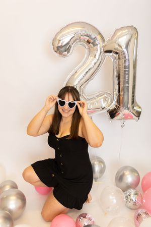A celebratory scene with silver number balloons, pink and silver party decorations, and someone in black posing against a white wall.
