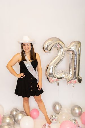 A celebratory photo shoot with silver balloon numbers, pink and silver balloons, and a black dress against a white backdrop.