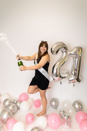 Woman in black dress poses with silver number balloons and pink and silver decorative balloons against white wall.