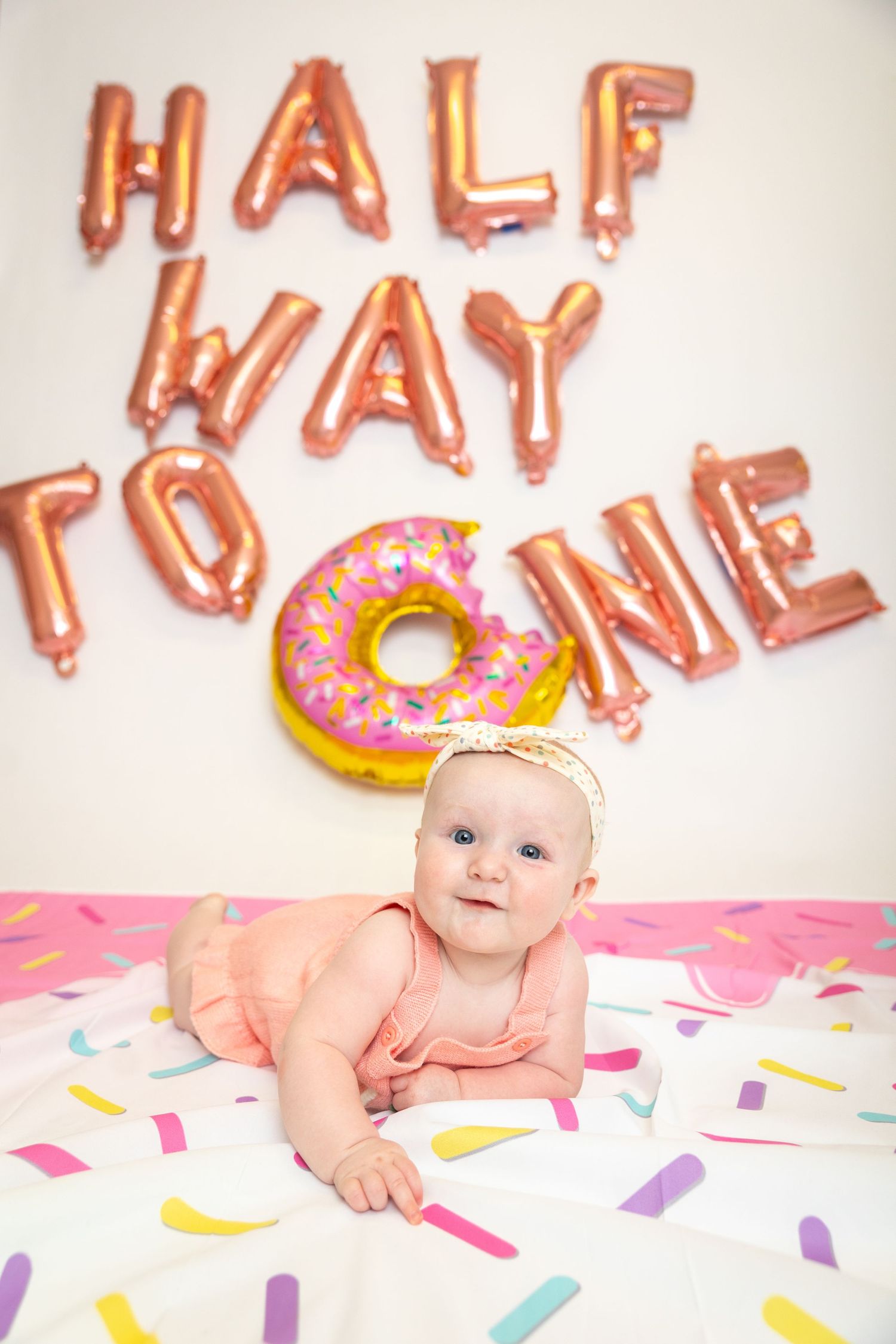 Colorful 'Half Way to One' balloon letters and donut decoration create festive six-month milestone celebration scene.