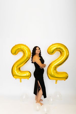 A person in a black dress poses with large gold metallic number 2022 balloons against a white backdrop.