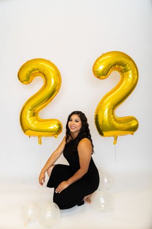 Gold metallic number 22 balloons float behind a figure in a black dress posing against a white backdrop.