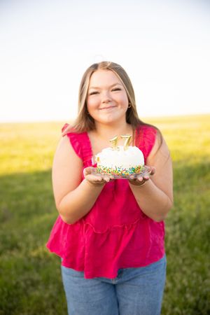 Person in red ruffled top holds slice of white cake against sunny outdoor backdrop.