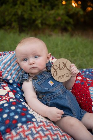 Baby in denim overalls lying on red, white and blue quilt with 6 month milestone marker.