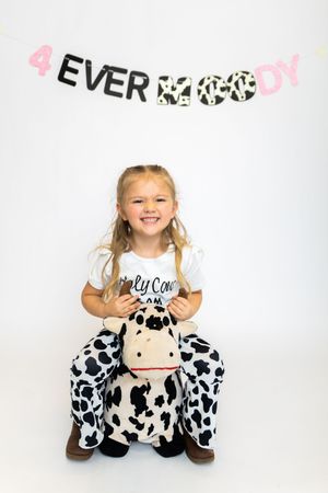 Young girl in white shirt poses playfully with black and white spotted cow plush toys against white backdrop.