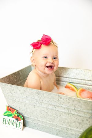 A baby wearing a pink bow smiles joyfully while sitting in a rustic metal container with summer-themed decorations.