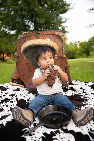 A small child sits on a black and white cowhide blanket wearing blue jeans and a cowboy hat in a grassy field.