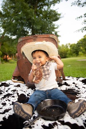 A small child wearing a cowboy hat sits on a cowhide blanket outdoors in a park setting.