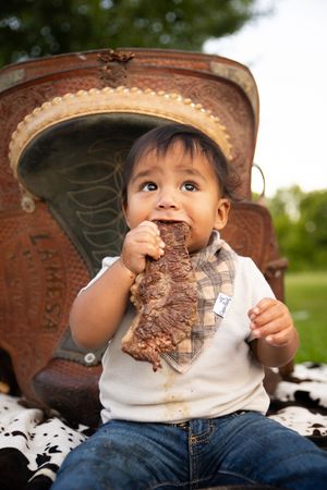 A young child in a cowboy hat enjoys eating an ice cream sandwich outdoors on a sunny day.