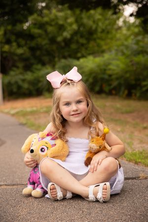 A child sits on a path outdoors with stuffed animal toys and wearing a pink bow in their hair.