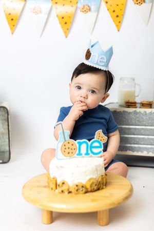 Baby in blue outfit sits at wooden table with cookie-themed party decorations and yellow banner hanging above.