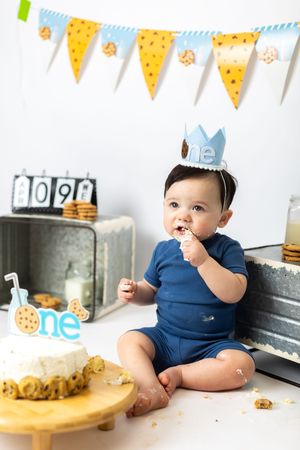 A baby in a navy blue onesie sits on the floor during a first birthday cake smash photo session with festive decorations.