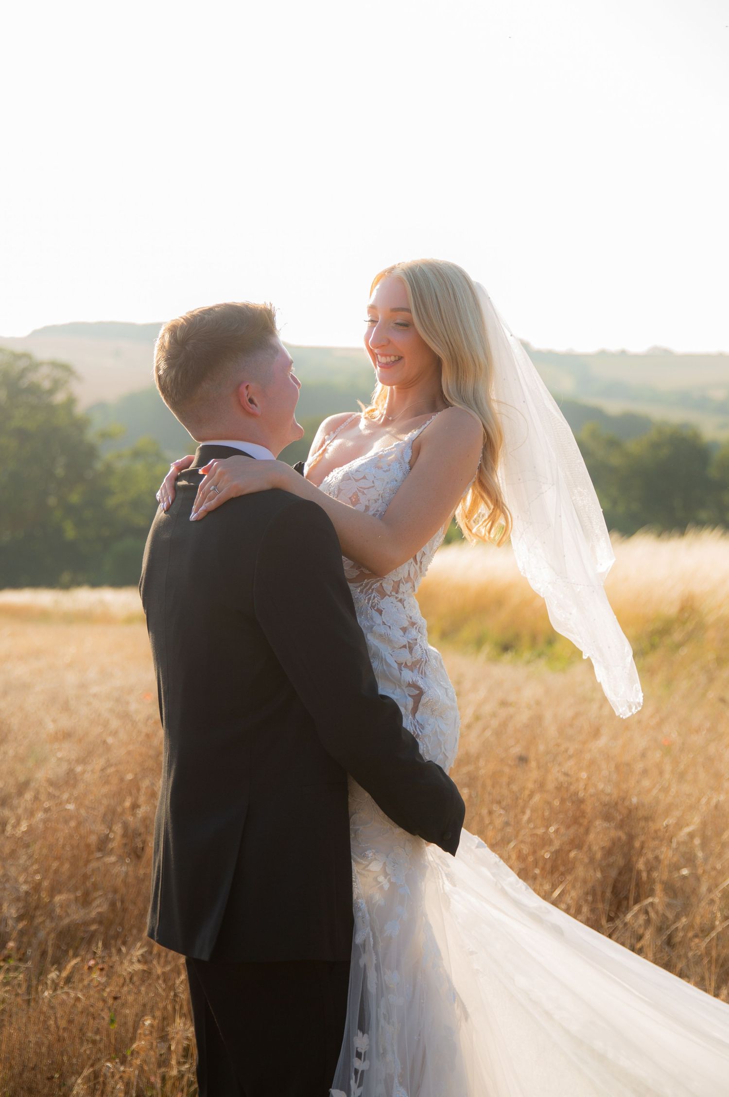 A couple in wedding attire embraces in a sunlit wheat field as the bride's veil flows in the breeze.